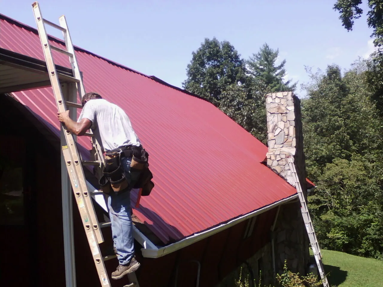 Roofer on a ladder beside a red metal roof and stone chimney — Soffit & Fascia Repair by Roofing Brothers in Elk Mountain